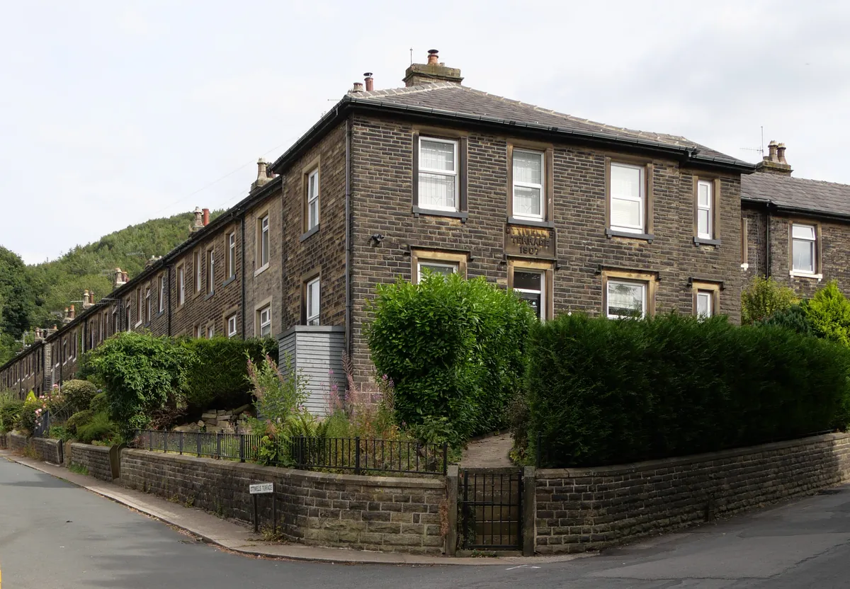 A long row of dark stone terraced houses on Ottiwell Terrace in Marsden, with wooded hills behind